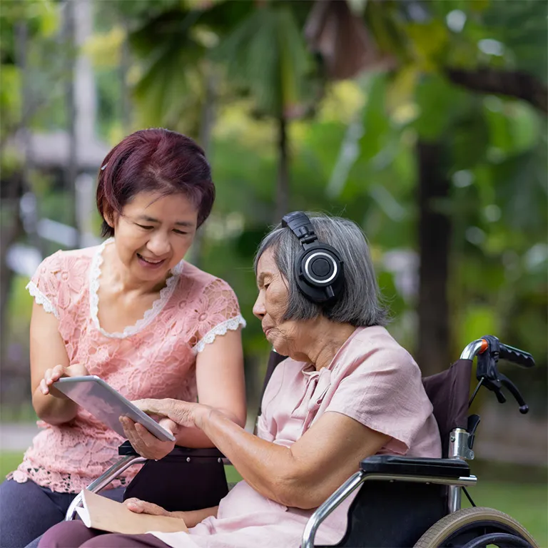 Elderly woman in a wheelchair wearing headphones and listening to music while another woman shows her a tablet, in a green outdoor setting